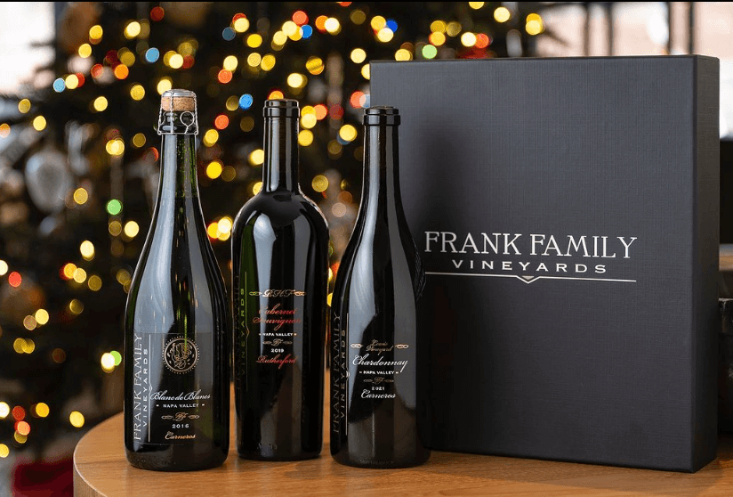 A picture of 3 bottles of Frank Family vineyards wine brand on a table. Beside the bottles are an elegant box case with the wines logo. The background consists of holiday lights and a Christmas tree.