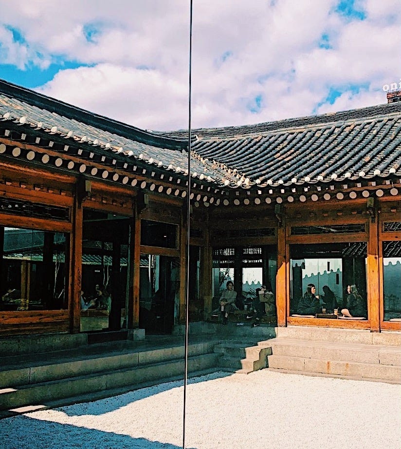 An traditional Korean house and roof, with a wooden structure and big windows, where you can see people sitting and chatting