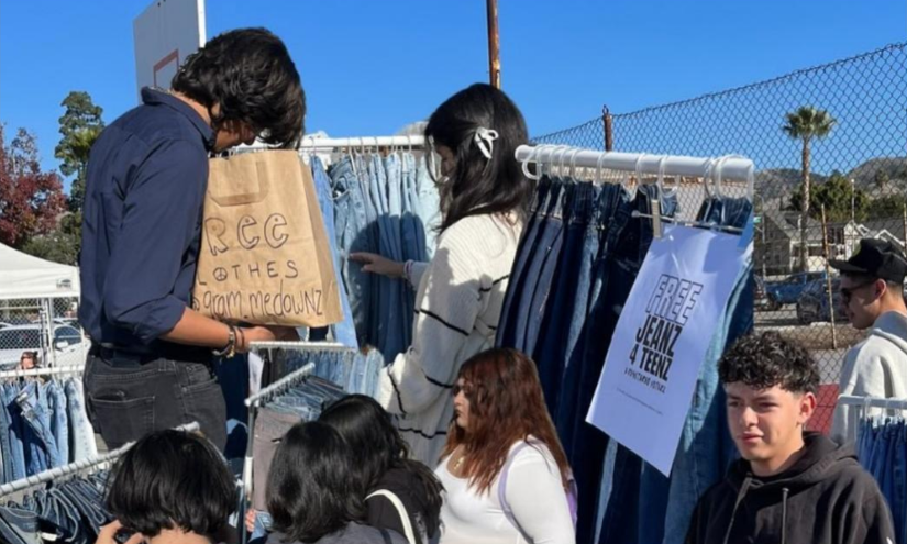 During a bright blue day, there are 7 people shopping. 2 are looking at a rack with one holding a bag reading “Free Clothes” in marker, the other four are on the bottom and are looking at clothes racks. There is a sign above one reading “Free Jeanz 4 Teenz