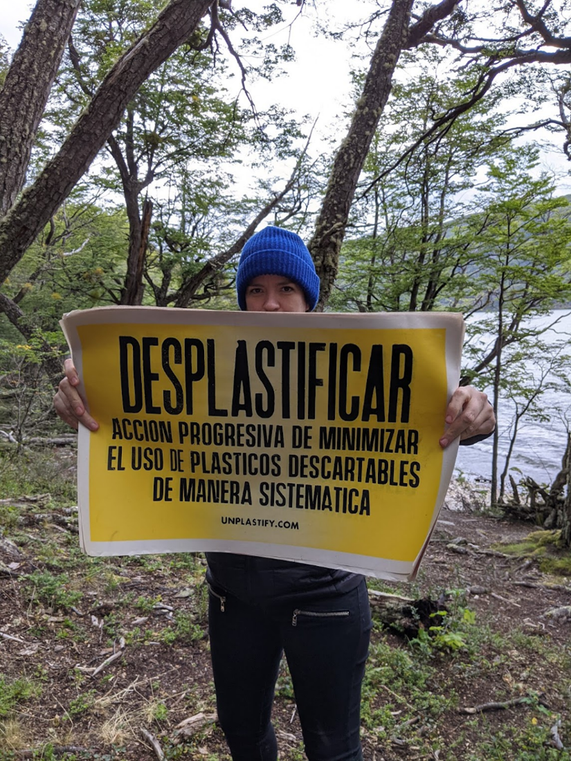 Person standing in forest in blue cap holding yellow sign with “Deplastify” instructions in Spanish.