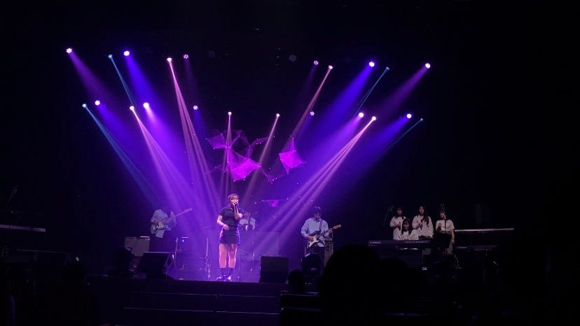 The photo shows a music performing stage and a female is singing in the middle. On the stage, there are also a few people playing instruments like guitar/drum/piano and a chorus. In a huge projection screen behind the singer, constellation-like particles and shapes are drawn in pink color. The stage is full of purple, pink, and blue lighting. Most of the lighting is pointing at the singer.