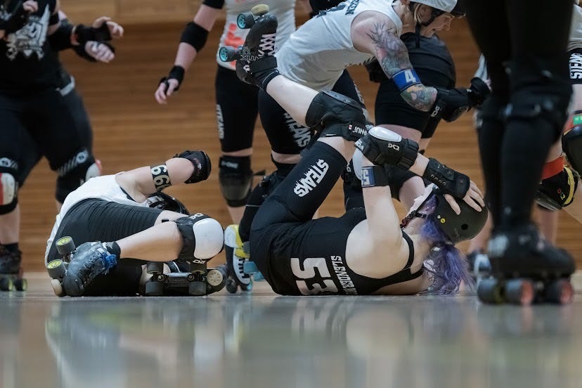 Professional sports photo of me lying on my back on the floor in a messy, tangled up roller derby game situation.