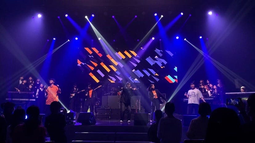 The photo shows a music performing stage and a group of vocalists singing in the front. On the stage, there are many people playing instruments like guitar, drums, piano and two chorus groups are on both sides of the stage. A huge projection screen behind the singer shows animated geometric shapes of irregular rectangles and trapezoids. They are all tilted about 45 degrees and colored in orange, yellow, purple, and more.