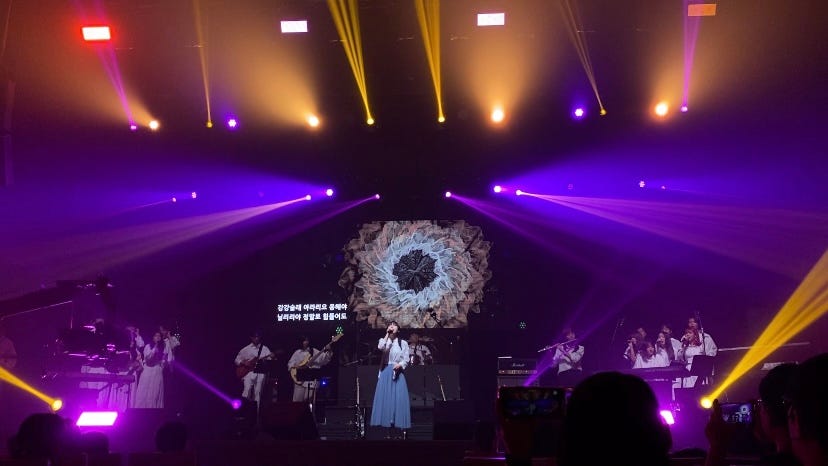 The photo shows a music performing stage. A woman is singing in the middle of the stage, two men are playing guitars on her right, and a woman playing flute on her left. Two chorus groups are singing at both ends of the stage. In a huge projection screen behind the singer, flower-like shaped perlin noise lines are drawn. The stage is full of purple, pink, and yellow lighting.