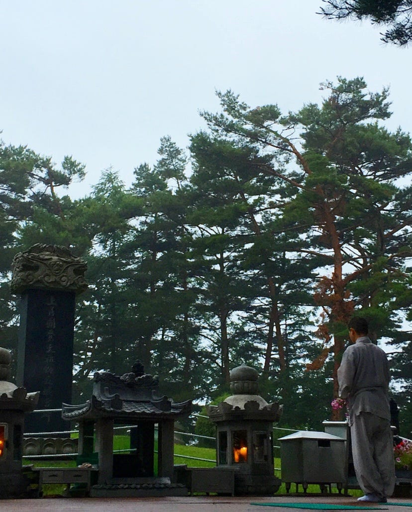 A buddhist monk praying deep in the mountains. There are statues that he is standing in front of. He is surrounded by trees.