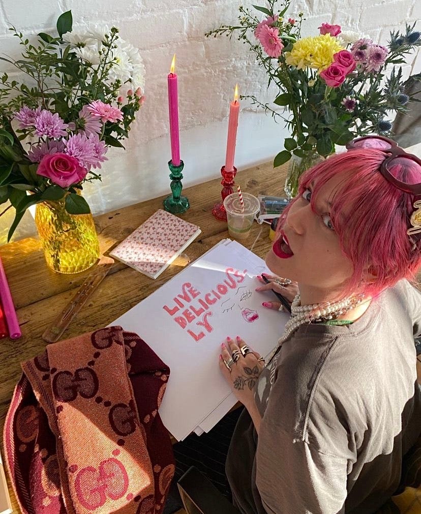 Florence Given sat at desk drawing a ‘live deliciously’ illustration with colourful candles and flowers around her