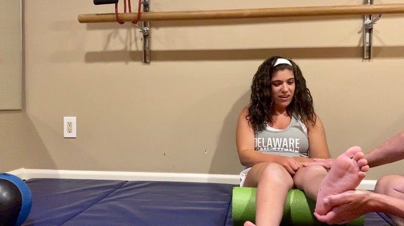 Julia sits on the mat while her physical therapist stretches her muscles.