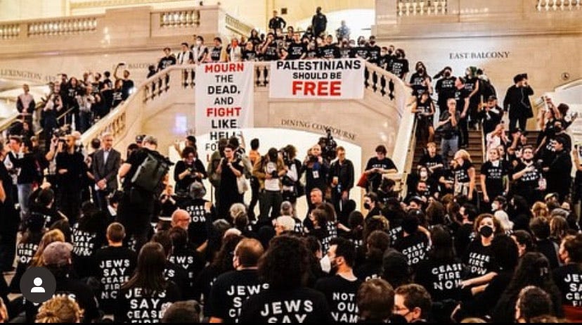 image showing a large crowd (perhaps hundreds) of people in black t-shirts saying “Jews say ceasefire now” crowding around a stairway at Grand Central station in NYC with two signs in red and black lettering draped over the railing. One reads “Mourn the dead, fight like hell for the living”, the other reads “Palestinians should be free”