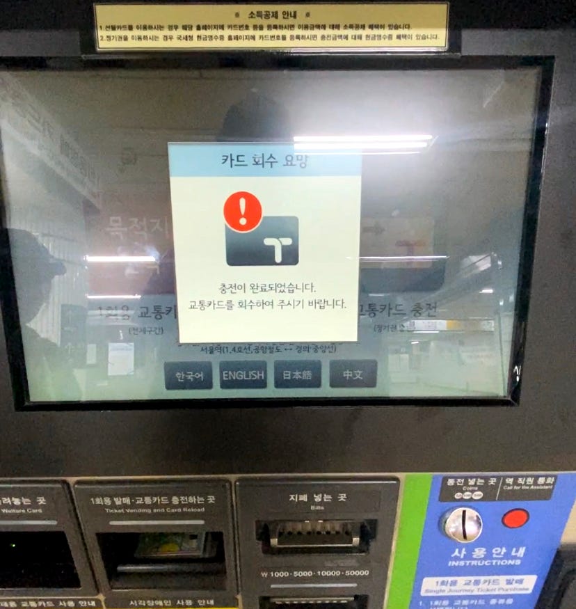 Tela de um totem de carregamento de metrô. Há uma janela aberta na tela com um desenho de um cartão e um ponto de exclamação vermelho. Uma mensagem abaixo em coreano que diz que o cartão de transporte está carregado.