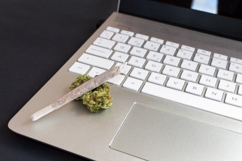 A cannabis joint on top of a dried cannabis flower sitting on top of a silver laptop computer.