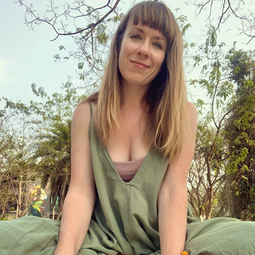 A photo of Andrea, sat cross-legged in a green all-in-one that matches the trees in the background. Sending you empathy from an open heart.