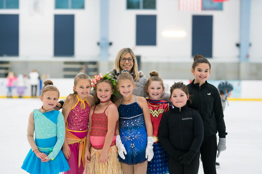 Martine De La Torre poses with a group of young skaters on the ice. Some are wearing colorful skating costumes and others are wearing black jackets and leggings. De La Torre stands behind the skaters smiling. She is a white woman with shoulder-length blonde hair and glasses.