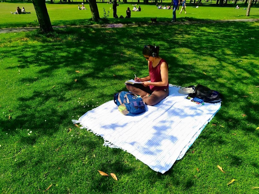 Foto tirada da perspectiva de uma pessoa em pé de uma mulher branca, jovem, de camiseta vinho e calça marrom de moletom, sentada sobre um plano branco num gramado verde, escrevendo à mão num caderno. Ela está de headphone, com uma mochila azul diante das pernas cruzadas, ladeada por outra mochila. Há árvores e pessoas ao fundo, e algumas folhas secas espalhadas pelo gramado.