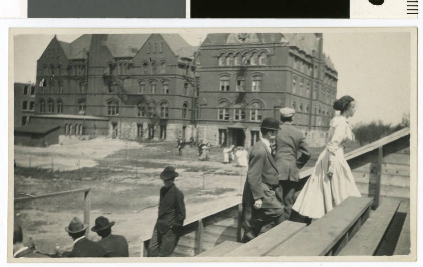 People climb the athletic bleachers in the foreground. In the background are the East and West Wings of Old Main.