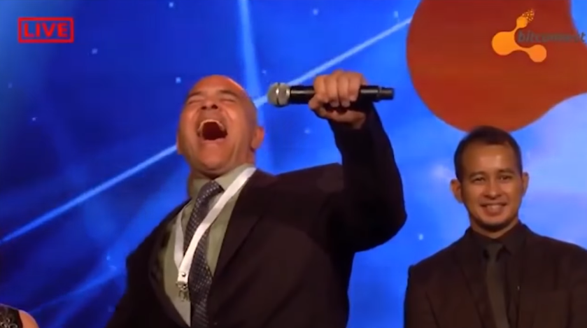 A picture of Carlos Matos, infamous BitConnect promoter, singing into the microphone on stage at a BitConnect event.