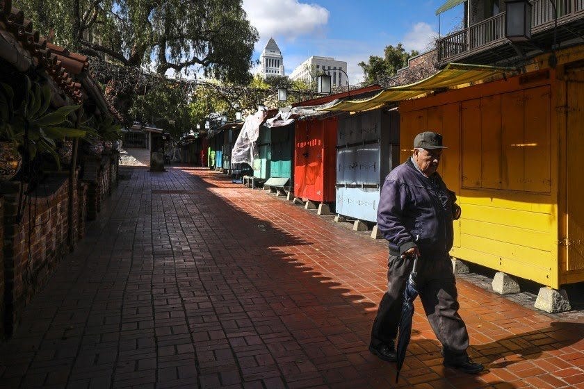 A lone man walks past turquoise, orange, blue, and yellow stalls. The walkway is empty and all the stalls are shuttered.