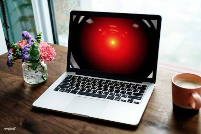 A laptop on a desk; the laptop’s screen is filled with the glowing red eye of HAL 9000 from 2001. Image: Cryteria (modified) https://commons.wikimedia.org/wiki/File:HAL9000.svg CC BY: https://creativecommons.org/licenses/by/3.0/deed.en