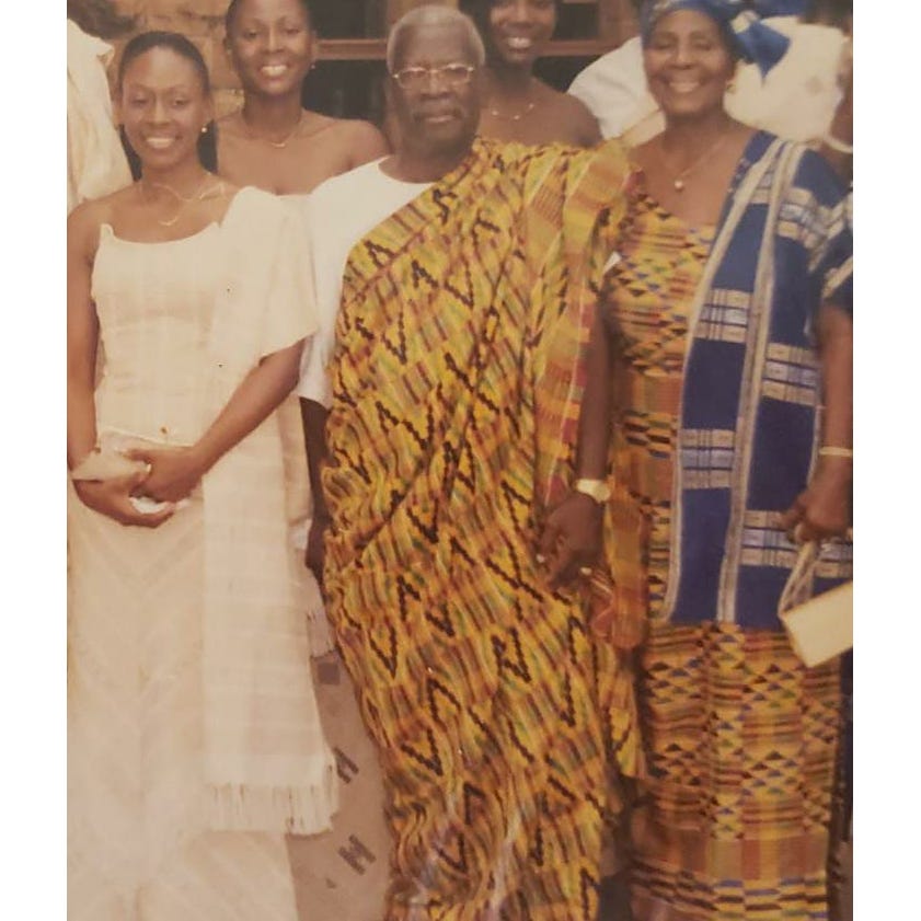 A young Adoley in a white dress smiles while standing next to her proud father and mother, both of whom are formally dressed in traditional Ghanian attire.