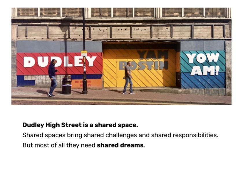 Signage art installation on a boarded up retail unit on Dudley High Street created as part of Do Fest 2019. The multicoloured sign reads: Dudley Yam Bostin Yow AM! Below the photo reads the words: “Dudley High St is a shared space. Shared spaces bring shared challenges and shared responsibilities. But most of all they need shared dreams.”