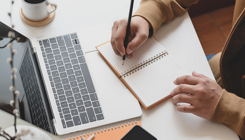 hands writing in a notebook sitting on top of an open laptop