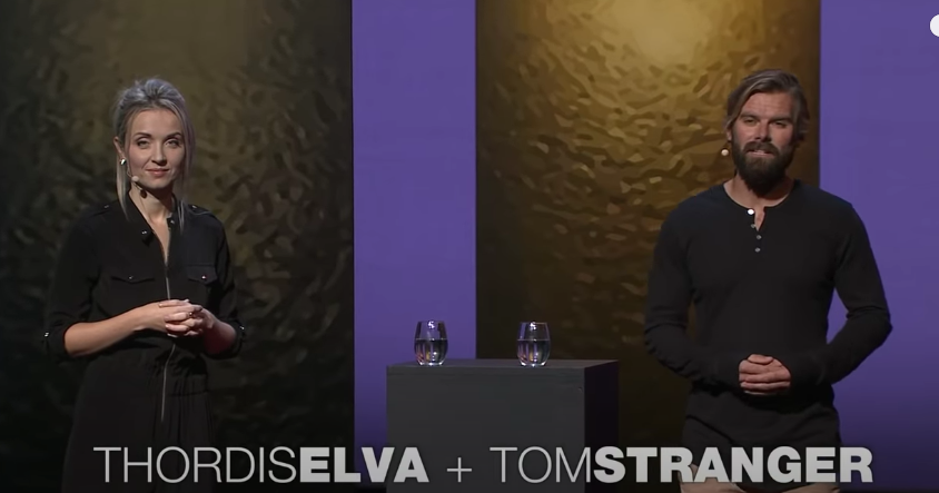 Thordis and Tom both dressed in black stand 6 ft apart in front of copper panels with purple panels between, black table between them with water glasses