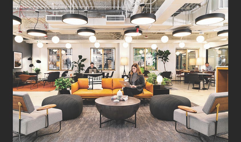 In a large office with tall ceilings and overhead large lights, there sits a girl on a medium sized orange leather couch. She is wearing professional/comfortable clothes as she works from her laptop in a shared co-working space. There are a few others in the room also working on their laptops.