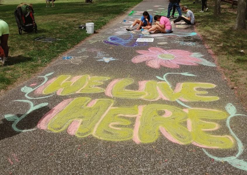 A sidewalk chalk drawing that says “WE LIVE HERE.” At the top of the sidewalk/photo is a group of children drawing with the chalk.
