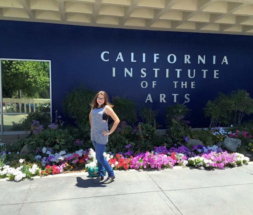 A teenage girl standing in front of a blue wall with “California Institute of the Arts” written and a flower garden below.