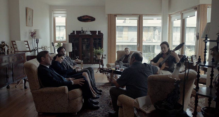 Six professionally dressed people sit in a living room. Two play the guitar. Colin Farrell and Lea Seydoux are central.