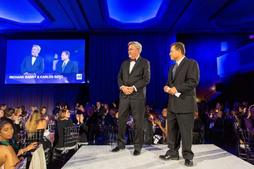 Rich Barry and Carlos Soto stand on a gray stage, with blue lights on the ceiling, and their faces up on a big screen behind them. Tables of people surround them as they watch the gentlemen speak.