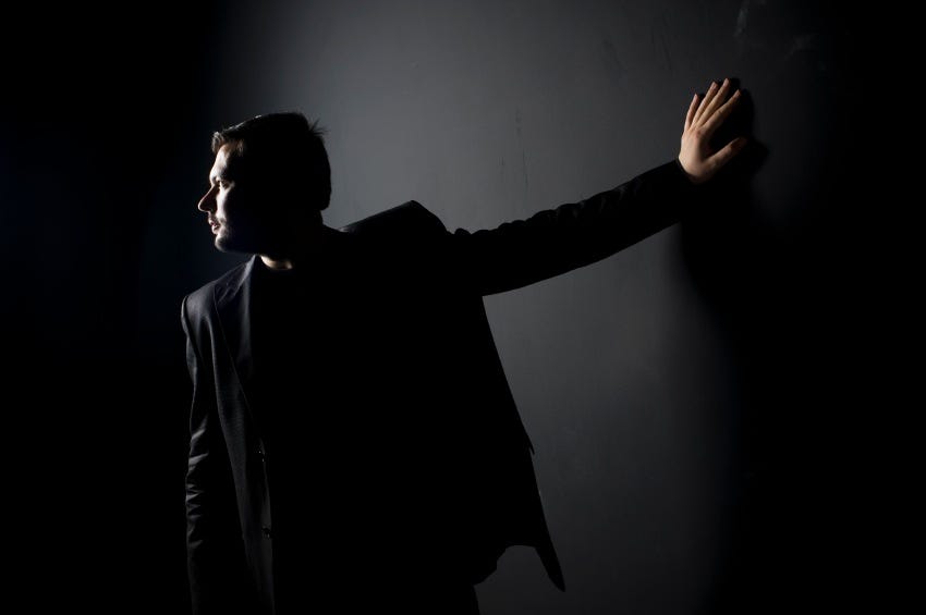 Image: A man in dark clothes and dim light, hand against the wall and looking the other way.