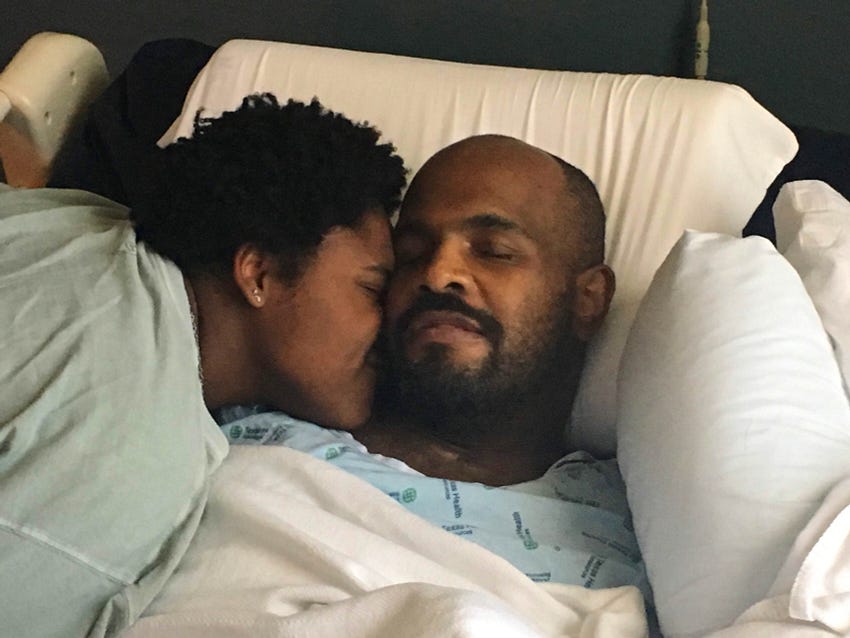 A couple is laying together on a hospital bed. The woman is nestled in to the man’s neck. Both of their eyes are closed.