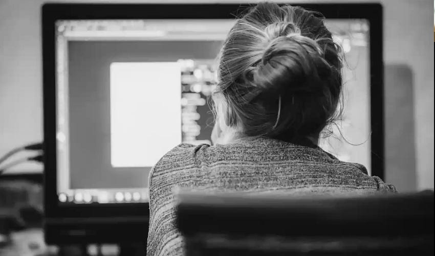 Woman with blonde hair in a bun facing away toward a computer screen.