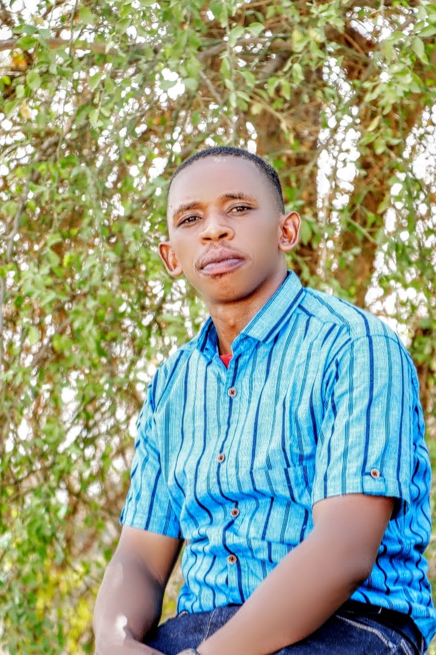 Mr. Kabagamba Byaruhanga Nestori sitting down in front of trees, looking at the camera. He wears a blue colar shirt and jeans.