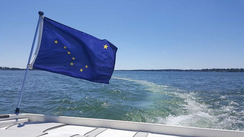 The Alaska State flag flying from the stern of a ski boat, on a large lake, with a blue green wake following behind.