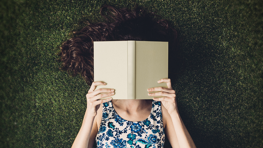 A woman laying on the grass with a book in front of her face for A Writing Habit That Doubles Reader Engagement