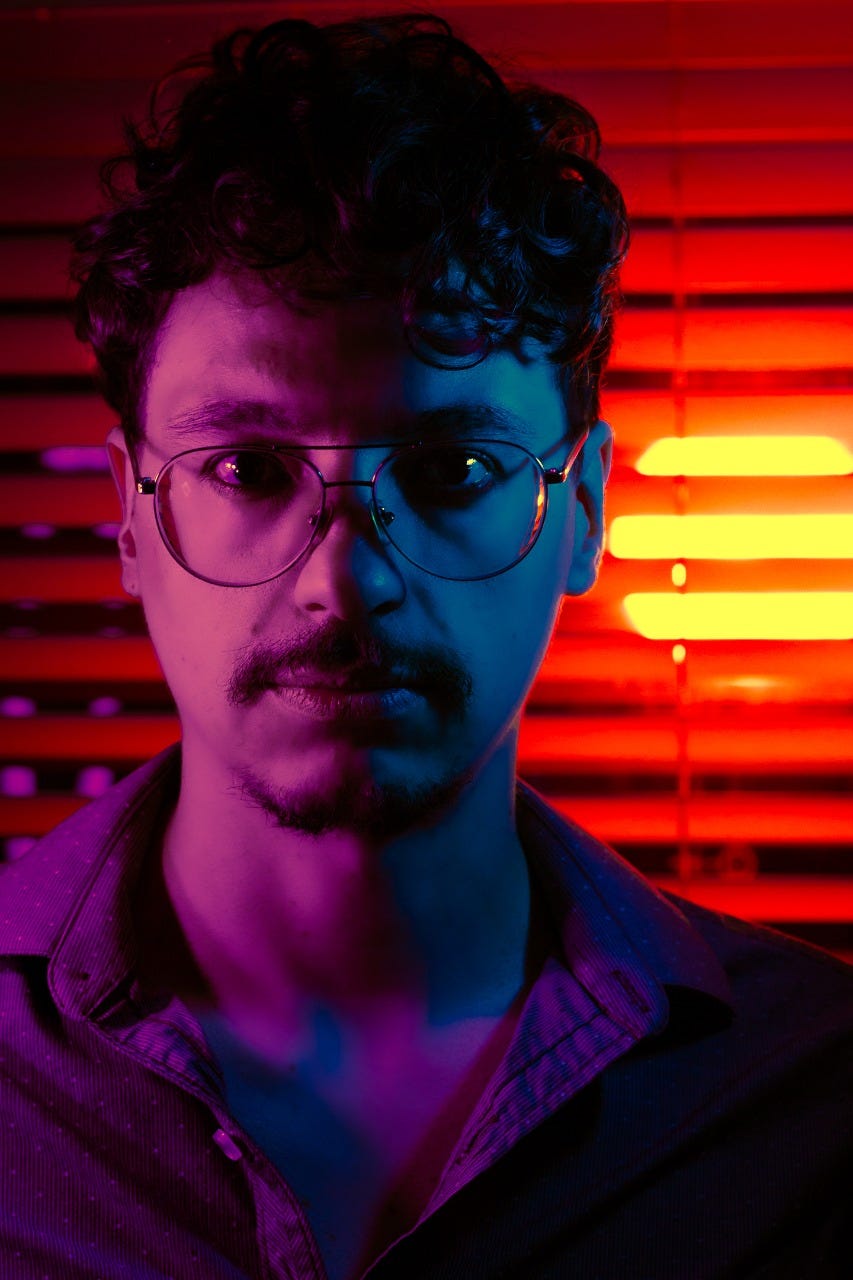 A portrait of the author looking straight at the camera. The left side of his face is lit in pink while the right side is lit in blue. In the background, a circular orange light pierces through horizontal blinds.