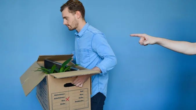 Photo of man in a blue dress shirt carrying a cardboard box full of items from an office. Someone else’s hand making a gesture indicating “get out” is visible on the right side of the photo.