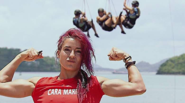 A proud and confident Cara Maria stands in front of a scenic water backdrop, flexing her muscles in a triumphant pose after a daily challenge win on “The Challenge.” She’s wearing a red tank top emblazoned with her name, and her wet crimson hair is a testament to the intensity of the competition. Behind her, other competitors are suspended in mid-air, adding to the dramatic context of her victory.