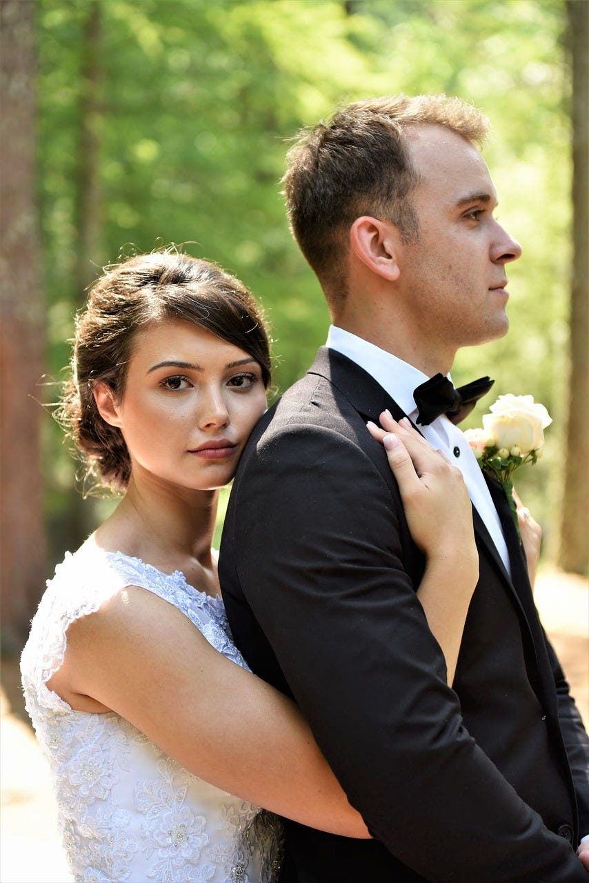 Image of a couple on their wedding day — the bride is a brunette in a white dress and she stands behind the groom, resting her cheek on his shoulder and her hand on his chest. He has short hair and wears a dark suit and bow tie with a white rose in his buttonhole