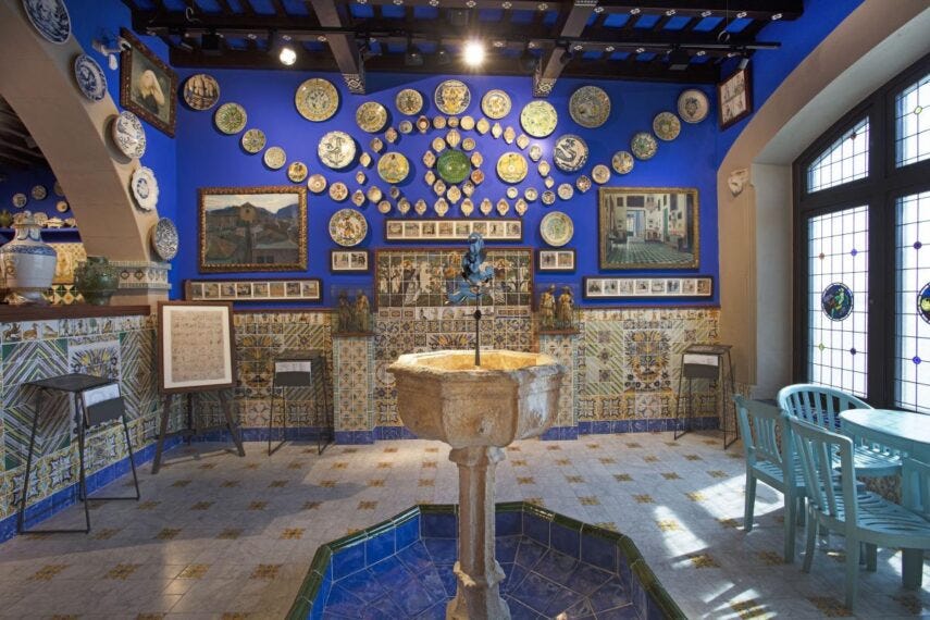 A photograph of Cau Ferrat, the home and studio of Santiago Rusinol in the town of Sitges, Spain, shows a fountain court with blue walls decorated with ceramic plates.