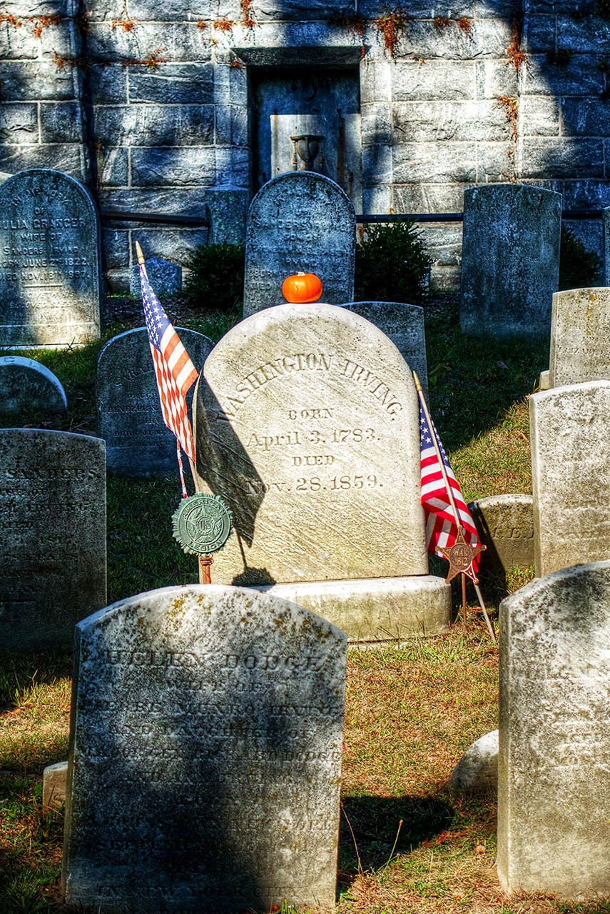 A Halloween decoration, a pumpkin, sits on top of a tombstone inscribed with the name “Washington Irving” in a cemetery.