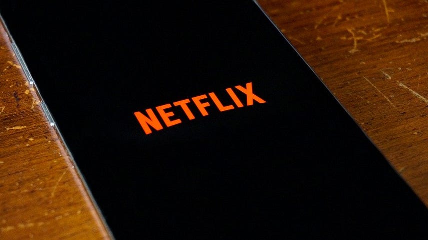 Imagem de um celular em cima de uma mesa de madeira. O celular estar com a tela na cor preta e no centro dela a palavra Netflix é exibida.