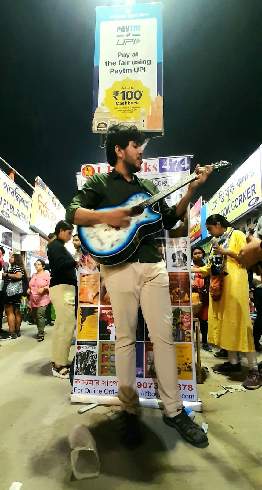 A boy with a guitar in hand singing country roads by Pete Seeger standing in front of a poster inside Kolkata Book Fair 2023