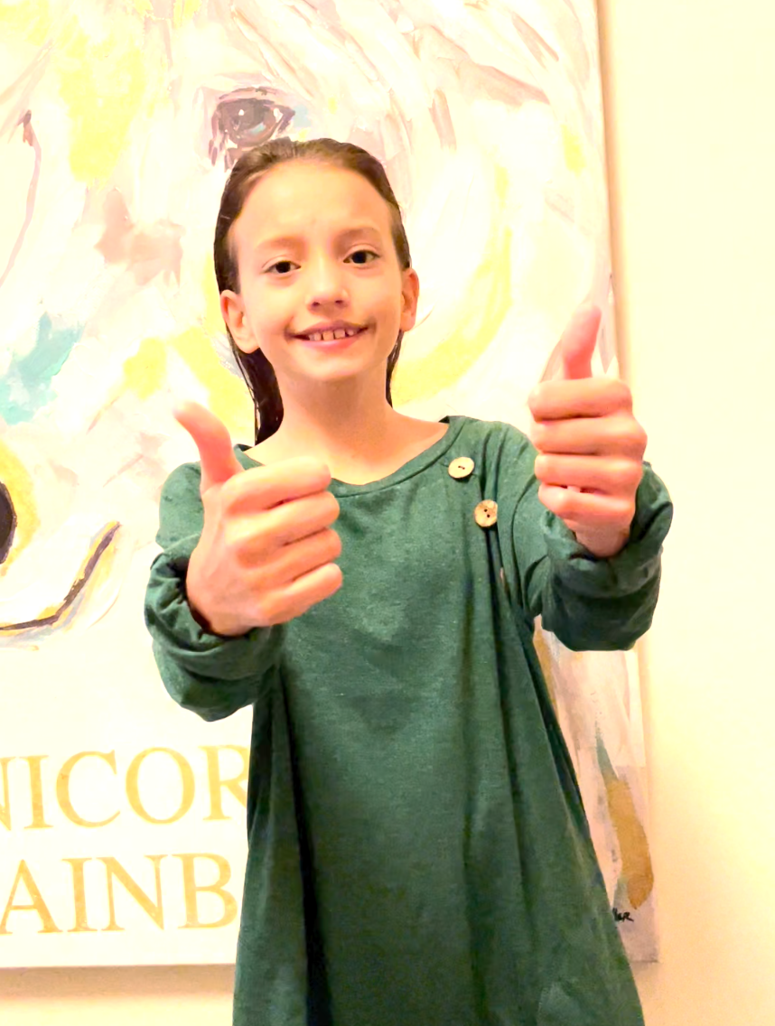 A picture of the writer’s daughter, Sofia, who is twelve year old. Sofia is smiling and is giving two thumbs up. She has long brown hair and is wearing a green sweatshirt dress to celebrate World Cerebral Palsy Day, on October 6th.