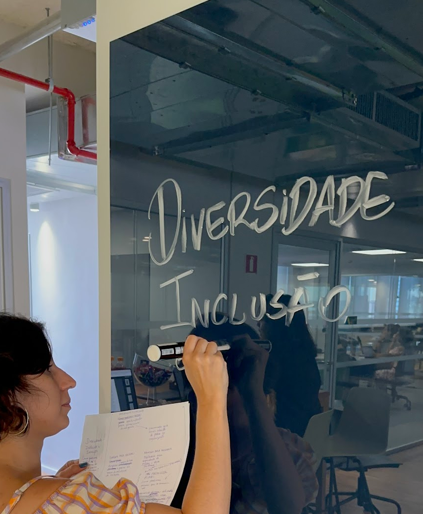 Vanessa, mulher branca, de cabelos curtos pretos e baixa estatura escreve no quadro de vidro com uma caneta de tinta branca as palavras "diversidade" e "inclusão"