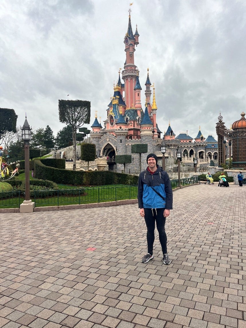 Me in a two-tone blue coat and beanie hat, stood in front of Sleeping Beauty’s castle in Disneyland Paris’ Magic Kingdom