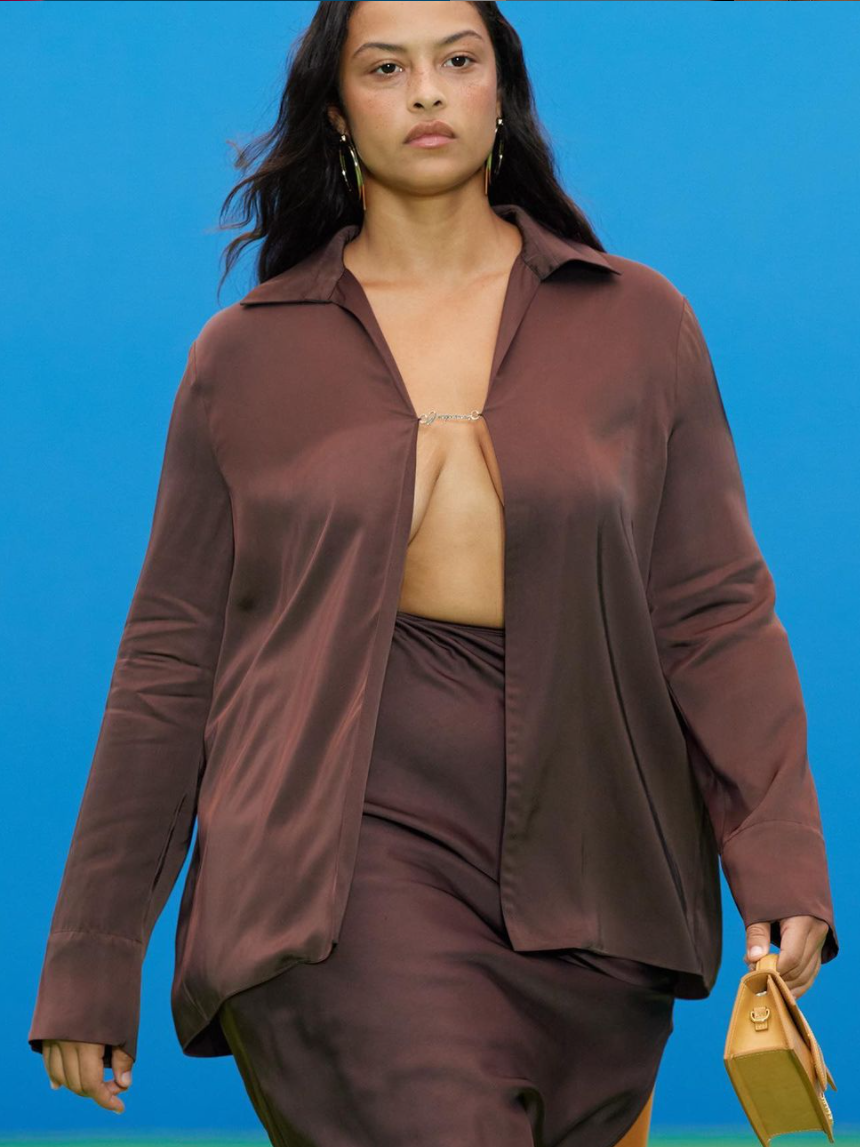 Model wearing brown top and brown skirt with a blue background. Model has a serious face.