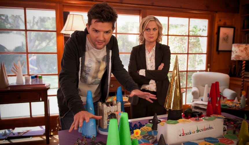 A still frame from Parks and Recreation showing Adam Scott’s character explaining a complicated board game; Amy Poehler stand behind him, skeptical