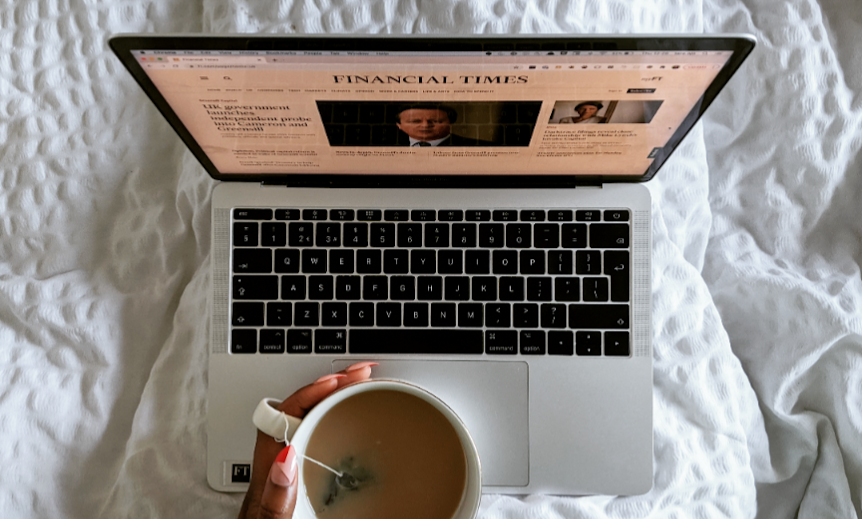 Laptop showing the Financial Times homepage on a bed with a cup of tea in hand and funky red and pink nails